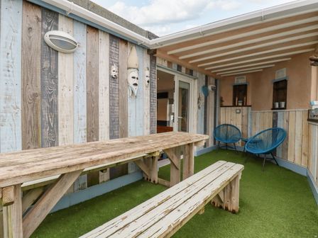 An outdoor covered seating area with a wooden picnic table and bench two blue chairs and wood paneling on the walls at Sailor's Rest in Newquay