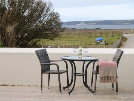 A patio with a round glass table and two wicker chairs with a bottle of wine and two glasses with a blanket on one chair with a view of the sea at Beachcomber in Marazion