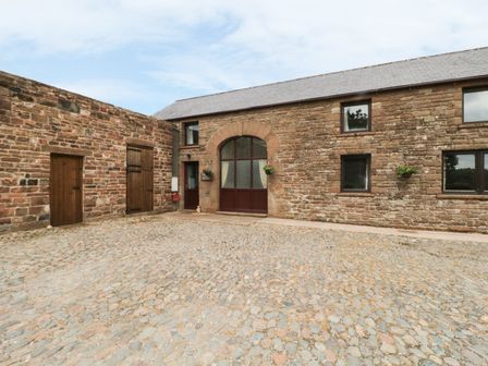 A stone building with wooden doors and windows and a cobblestone courtyard at Woodhead Farm Cottage in Penrith