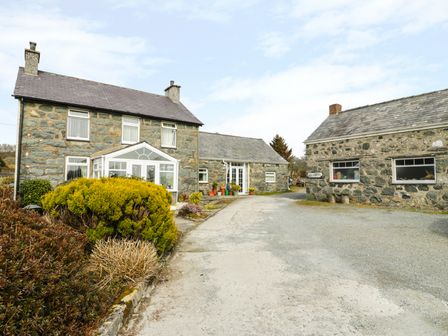 An exterior view of two stone buildings with gravel driveway and shrubs at Bwthyn Ael Y Bryn in Criccieth