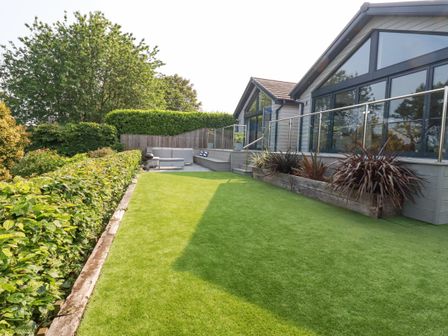 A backyard with green grass hedge and modern house with large windows and outdoor seating at Trenant View in Wadebridge