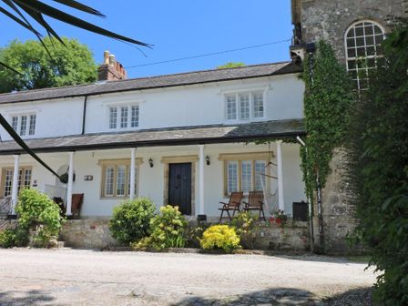 A white cottage with shuttered windows wooden chairs and green shrubs outside Sea Cliff Cottage in Pentewan