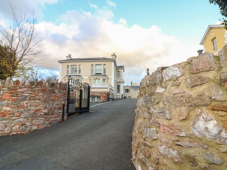 A driveway with a black gate leading to a beige house with multiple windows and stone walls on both sides at Cottage 1 Newcourt in Torquay