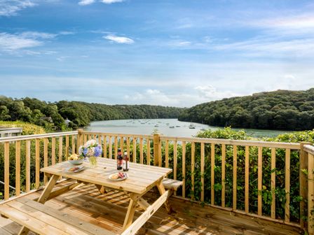 A wooden picnic table with food and drinks on a deck overlooking a river with boats and forested hills at Fal River Cottage in Malpas Cornwall