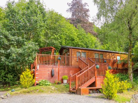A wooden cabin with a deck and stairs surrounded by trees and plants at Willow Lodge in Troutbeck Bridge