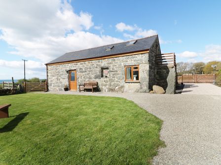 A stone house with wooden door and windows with a bench outside on a lawn with gravel path and a wooden fence at Sgubor in Chwilog