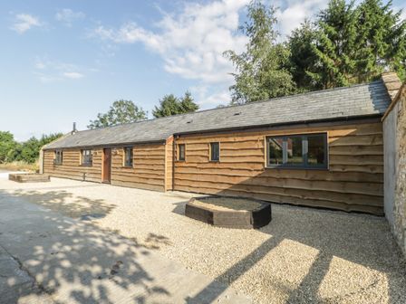 An exterior view of a single-story wooden building with gravel ground and trees in the background at The Milking Barn in Yeovil Somerset