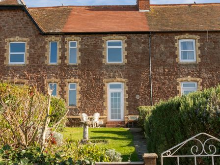 A garden with bushes and a bird bath in front of a brick house with multiple windows and a glass door at Harbour View Cottage in Watchet
