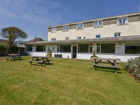 A grassy backyard with wooden picnic tables and a three story building in the background at 2 Brook Cottages in Hope Cove