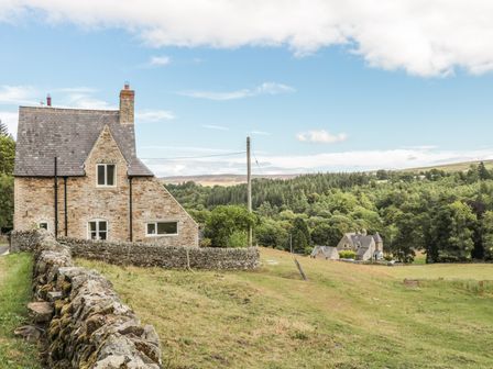 A stone house with a slate roof next to a stone wall in a rural area with trees and hills at Newbiggin Cottage in Blanchland