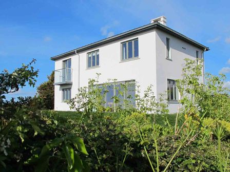 A two story white house with gray windows surrounded by green plants and trees at Kareena in Crantock