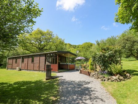 A wooden cabin with a covered porch and outdoor seating on a gravel path surrounded by trees and grass at Brook Lodge in Penrhiwllan