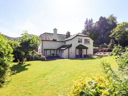 A white house with dark roof surrounded by a large green lawn and trees at Coed Derw Isaf in Betws-Y-Coed
