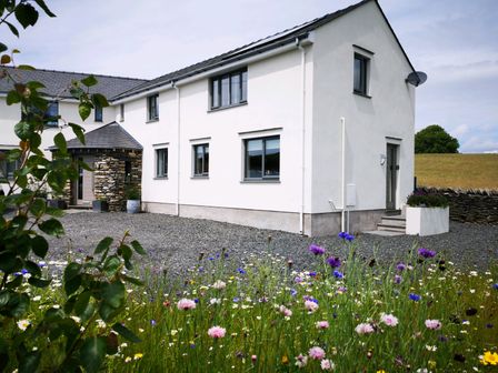 A white two-story house with a gravel driveway and wildflowers in the foreground at Yew Fold in Kirkby-In-Furness