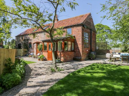 A garden with stone pathway and grass in front of a brick house with red roof tiles and wooden doors at The Stables in Roos