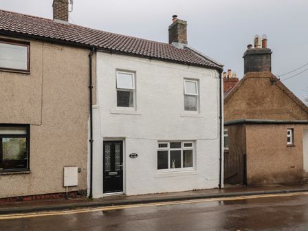 A white two-story cottage with a black door and three windows on a wet street at Magpie Cottage in Wooler