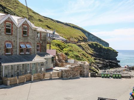 Stone buildings near a rocky coastline with green hills and the sea at Gull Rock in Trebarwith Strand