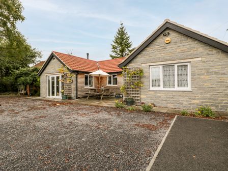 An exterior view of a single-story house with stone and wood siding a gravel driveway and outdoor seating with an umbrella at Vine Lodge Kingsbury Episcopi near Martock