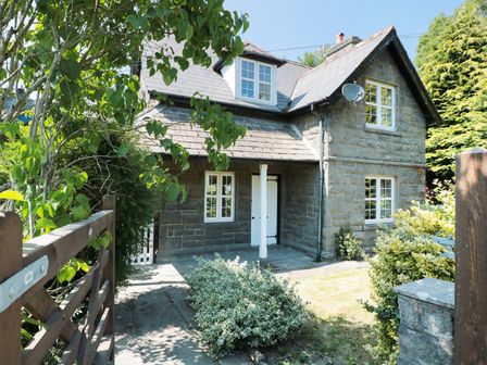 A stone house with white windows and a wooden gate surrounded by bushes at School House in Builth Wells