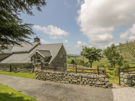 A stone cottage with a slate roof next to a stone wall and wooden fence with trees and hills in the background at The Stable Cottage in Llanbedr