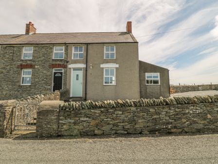 Exterior view of a stone and plastered house with two chimneys a stone wall and an iron gate at Glasfryn in Llanfaethlu