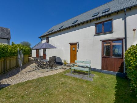 An outdoor patio area with a table and chairs under an umbrella and a wooden bench in front of a house at The Croft in Holsworthy