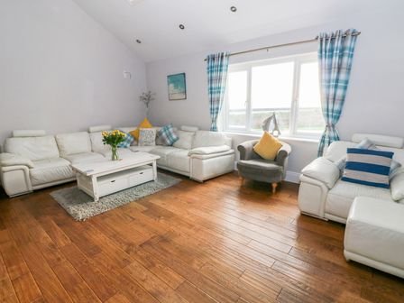 A living room with white sofas a gray armchair a white coffee table with flowers wooden flooring and blue check curtains at Swn y mor in Amroth