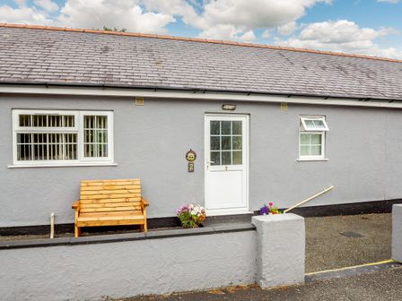 An exterior view of a grey building with two windows a white door a wooden bench and flower pots at 2 Black Horse Cottages in Pentraeth