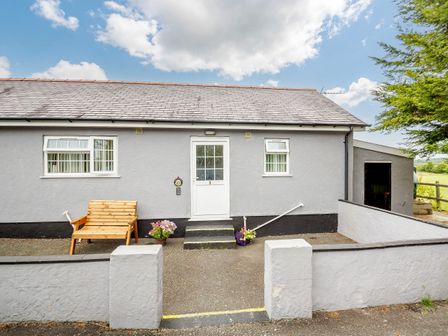 The front of a single-story grey house with a white door and two windows a wooden bench and flower pots outside at 3 Black Horse Cottages in Pentraeth