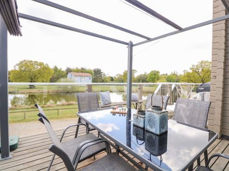 An outdoor deck with a glass table and chairs overlooking a pond with trees and a small blue house at Links View in Fritton