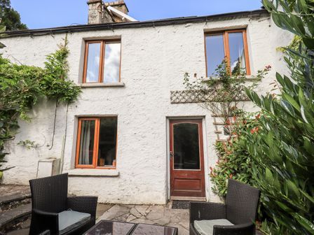 An outdoor patio area with two black wicker chairs and a table in front of a white stone building with four windows and a wooden door at 4 Greencross Cottages Burton-in-Kendal near Kirkby Lonsdale