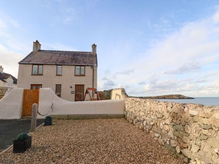 A two-story house with a gravel yard and a stone wall beside the sea at Beacon Cottage in Cemaes Bay