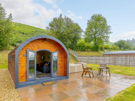A wooden pod with open doors on a tiled patio with two chairs and a small table at Corndon Pod in White Grit near Chirbury