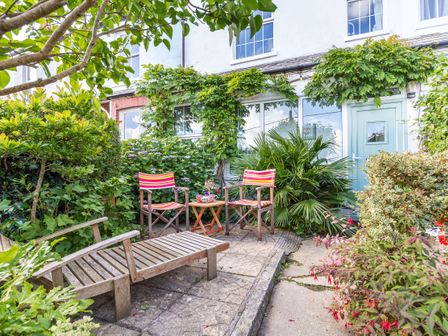 A garden seating area with wooden chairs and a small table surrounded by plants in front of a house with a light blue door at Downlea in Looe
