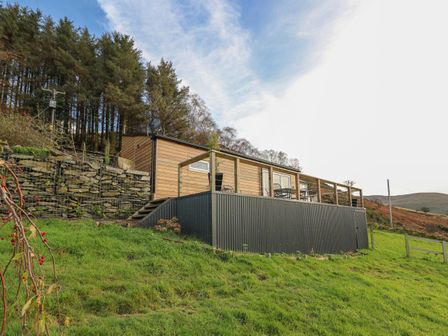 A cabin with a wooden deck and metal siding on a grassy hillside near trees at Haka Lodge in Devil's Bridge