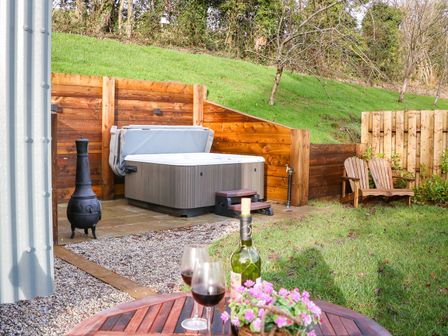 A backyard with a hot tub wooden bench a table with wine glasses and flowers and a metal chiminea at Little Silver Fox in High Bickington
