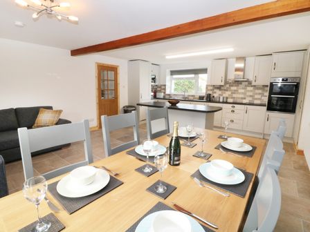 A dining table set with plates bowls wine glasses and cutlery next to a kitchen with cabinets an island and built in oven at Ysgubor Fawr in Llandwrog