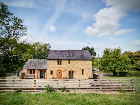 A stone house with a wooden fence and a patio with outdoor furniture surrounded by trees at Little Barn in Ascott near Chipping Norton