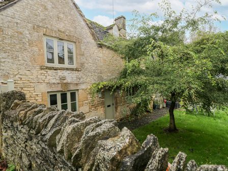 A stone cottage with green window frames and door behind a stone wall with a tree and grass in the garden at Tannery Cottage in Burford