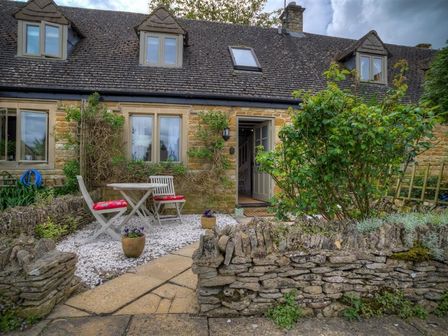 A stone cottage with a small patio table and two chairs with red cushions outside at Bobble Cottage in Little Rissington