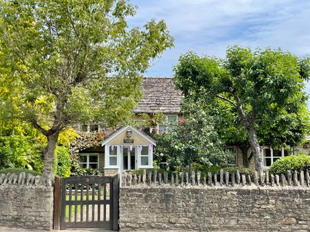 A cottage house with a wooden gate and stone wall surrounded by trees and plants