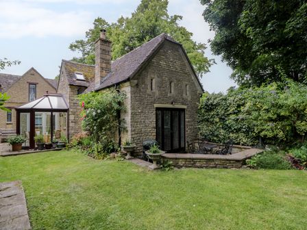 A stone house with a glass conservatory and a small patio with metal chairs and table in a garden