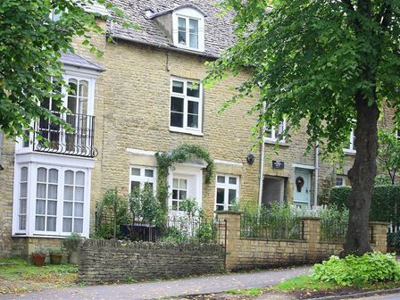 Stone houses with white windows and doors along a sidewalk with trees and greenery at Hare House in Chipping Norton
