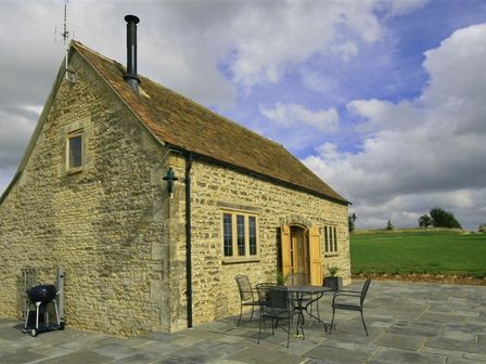 A stone cottage with wooden windows and door on a stone patio with metal table and chairs and a barbecue grill