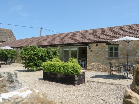 A stone building with a tiled roof and a courtyard with outdoor tables umbrellas and greenery at Nutchat Barn in Burford