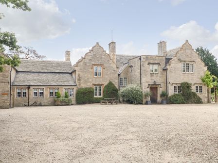 A stone house with multiple windows and gable roofs surrounded by gravel and bushes at Home Farm (16) in Witney