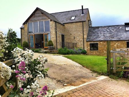 A stone house with large windows and a gravel driveway with flowers in the foreground at Stepwell in Moreton-In-Marsh