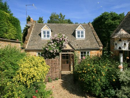 A cottage with stone walls and tiled roof surrounded by flowering bushes and a gravel pathway at Hadcroft Cottage in Aston Magna
