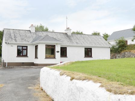 A white single-story house with a gray tiled roof and brown window frames next to a grassy area and a paved driveway at Daffodil Cottage in Grange County Sligo