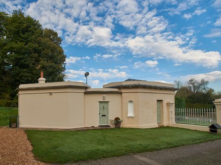A beige building with green doors and a grassy yard with tall trees and a partly cloudy sky in the background at West Lodge in Rockbourne